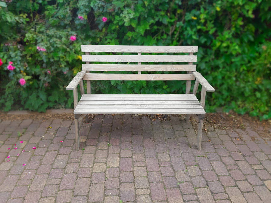 Grijze houten tuinbank voor 2 personen met armleuningen, geplaatst voor een groen bosje met roze bloemen op straatwerk.