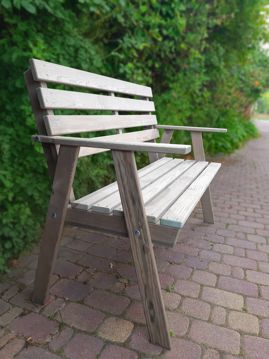 Grijze houten tuinbank voor 2 personen met armleuningen, schuin gefotografeerd, geplaatst voor een groen bosje op straatwerk.