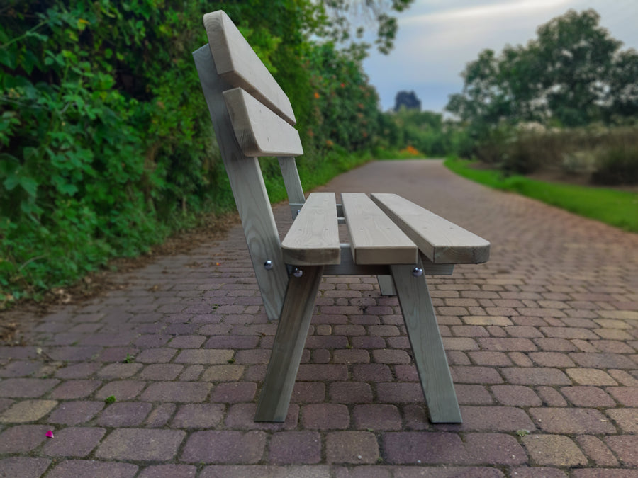 Grijze houten tuinbank voor 2 personen, schuin gefotografeerd, geplaatst voor een groen bosje op straatwerk.
