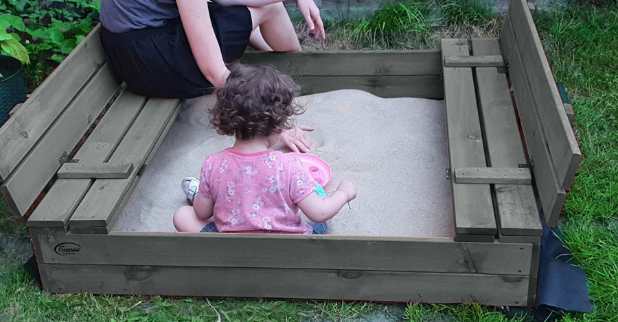 "Houten Grijze zandbak met deksel en bank, gevuld met zand, opengeklapte bankjes en een spelend kind."